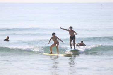 A man and woman riding surfboards on a small ocean wave during a sunny beach surfing lesson.