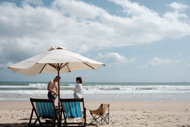 Couple relaxing on a sunny beach under a large white umbrella with blue beach chairs.