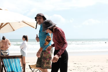 Two people, a man and a woman, walk along a sunny beach with an umbrella and ocean in the background, both wearing