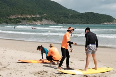 Instructors in orange shirts lead a group surf lesson on a sandy beach with ocean waves.