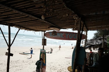 Tropical beach scene viewed from a rustic bamboo surf shack with people on the sandy shore and ocean waves.