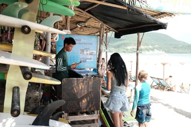 Surfers check in at a beachside surf school camp with surfboards stacked on a tropical shore.