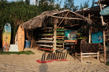 A rustic surf camp on a sandy beach featuring surfboards for rent and lessons under a thatched-roof hut, perfect for