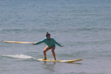 A female surfer in a blue long-sleeve rash guard balances on a yellow surfboard riding a small ocean wave.
