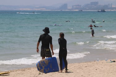 Two surfers standing on a sandy beach with a blue surfboard looking out at ocean waves.