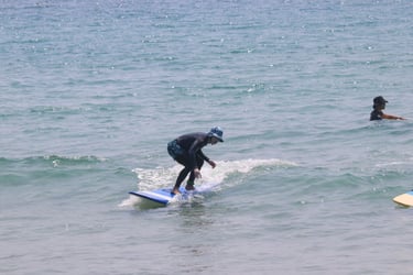 A person in a black wetsuit and blue hat surfs a small wave on a light blue surfboard in the ocean.