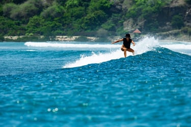A female surfer in a black swimsuit catches a blue wave near a tropical coastline.