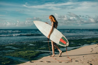 A red-haired female surfer carrying a white surfboard on a tropical beach at sunset.