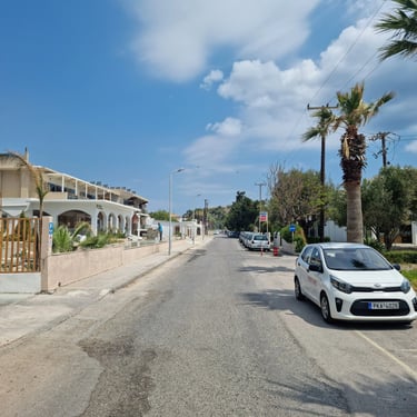 White Kia Picanto parked on a sunny street in Greece with palm trees and vacation apartments.