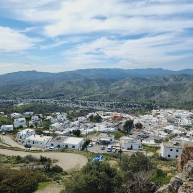 Panoramic view of white houses in a traditional Greek village nestled among rolling hills and mountains.