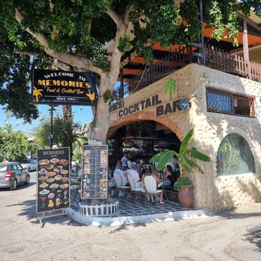 Memories Food & Cocktail Bar outdoor patio with guests, menu signs, and stone architecture under a large tree.