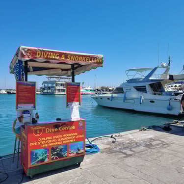 A scuba diving and snorkeling tour kiosk at a sunny Mediterranean harbor with luxury yachts in the background.