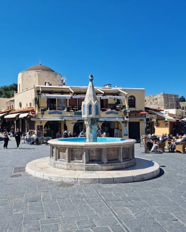 The ornate stone fountain in Hippocrates Square, Rhodes Old Town, with outdoor cafes and a mosque under a blue sky.