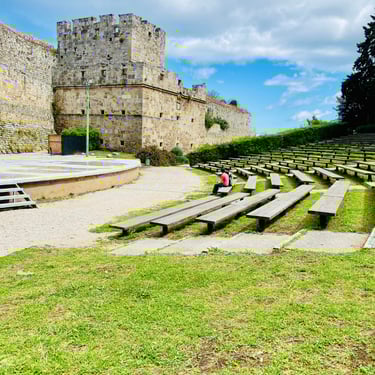 Outdoor stone theater with wooden benches next to the historic Palace of the Grand Master in Rhodes, Greece.