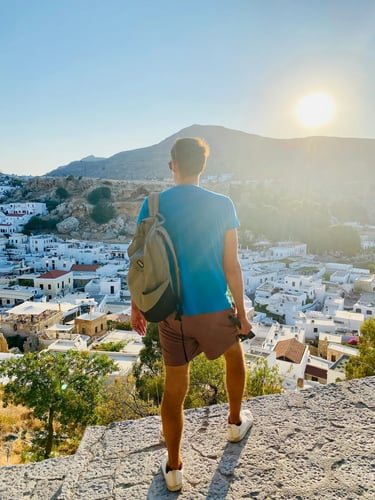 A male traveler with a backpack overlooks the white architecture of Lindos, Rhodes during a sunny Mediterranean sunset.