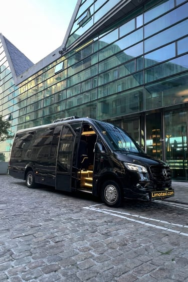 Black Mercedes Sprinter luxury limo bus parked on a cobblestone street in front of a modern glass building.