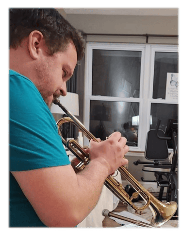 A man in a teal shirt plays a gold brass trumpet indoors near a window.