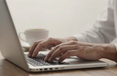 Close-up of elegant hands typing on a laptop, soft focus on a white ceramic cup of coffee, alabaster background.