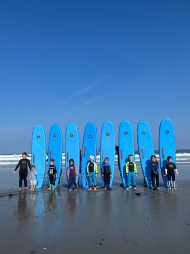 A group of children in wetsuits standing with blue foam surfboards at a kids surf camp on the beach.