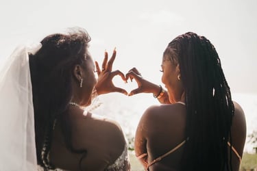 A bride and bridesmaid making a heart shape with their hands during a sunny outdoor wedding.