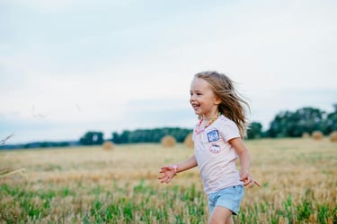 A happy young girl laughing and running through a golden wheat field with hay bales at sunset.