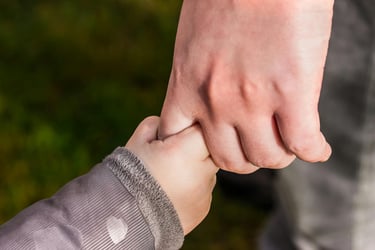 A close-up of a toddler holding an adult's finger while walking outdoors, symbolizing guidance and trust.