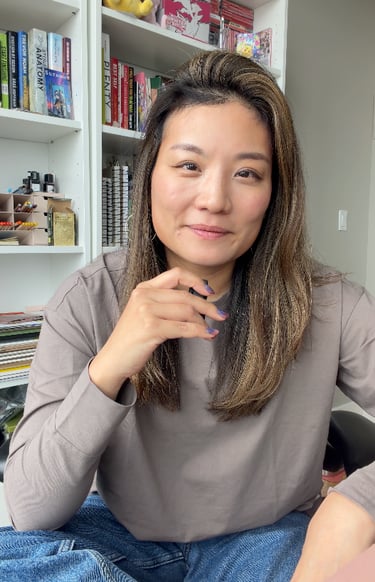 Smiling woman with highlighted hair sitting in front of a white bookshelf filled with books and art supplies.
