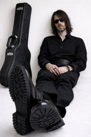 Trash Swallow of PreSexHeadache sitting in black clothing beside a guitar case