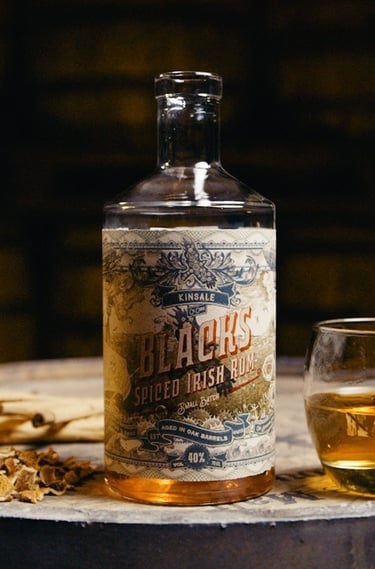 A bottle of Blacks Spiced Irish Rum on a wooden barrel with a glass of spirit and spices.