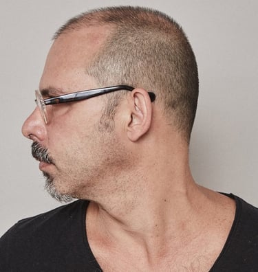 Side profile of a man with a buzzed haircut, goatee, and eyeglasses against a neutral background.