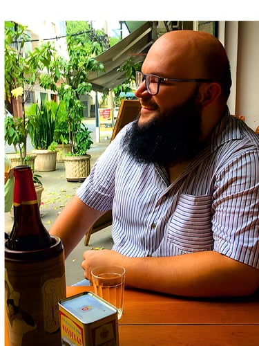 A smiling man with a full beard and glasses sitting at an outdoor cafe table with a beer.