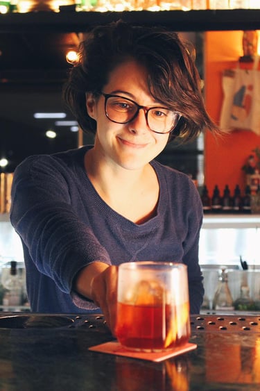 A smiling female bartender wearing glasses serves a craft cocktail in a glass at a bar.