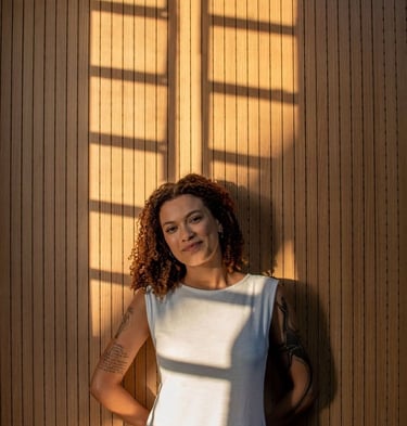 A smiling woman with curly hair stands against a wood-paneled wall with window shadows.
