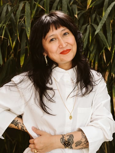 A smiling woman with dark hair and tattoos wearing a white shirt and gold jewelry against green leaves.