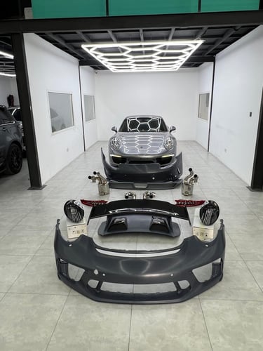 Gray Porsche 911 in a workshop with disassembled body kit parts, bumpers, and performance exhaust system laid out.