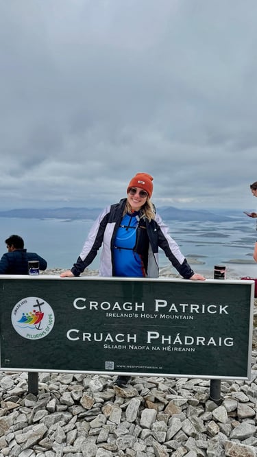 A smiling hiker at the summit of Croagh Patrick mountain in Ireland with scenic coastal views.