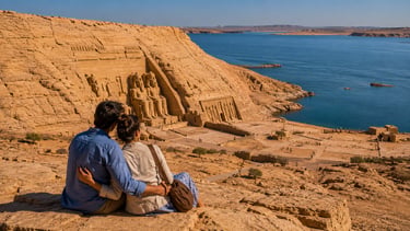 A couple overlooks the historic Abu Simbel temples and Lake Nasser in Egypt from a rocky desert cliff.