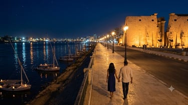 A couple walks along the Nile River at night, with the illuminated Luxor Temple and traditional sailboats in view.