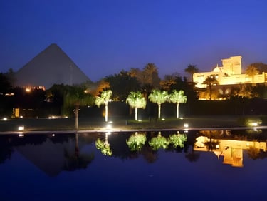 Night view of the Great Pyramid of Giza reflected in a luxury resort pool with palm trees.