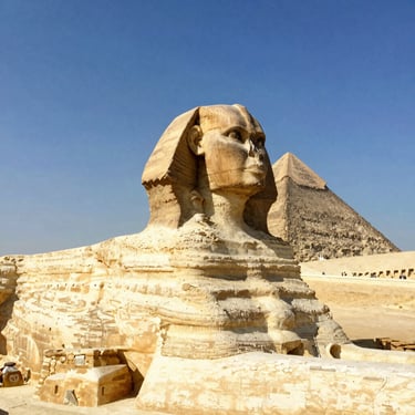 The Great Sphinx of Giza side profile with the Great Pyramid of Giza in the background under a blue sky.