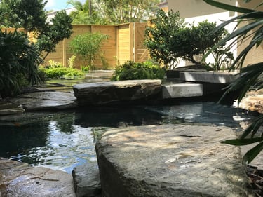 Serene outdoor pond with natural stone, lush green landscaping, wooden fence, and concrete steps, creating a tranquil garden