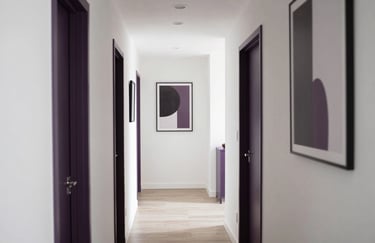 A bright photography shot of a communal hallway or transitional space in a modern home, featuring clean white walls, dark purple accents in the artwork, and soft, natural lighting.