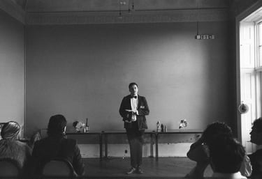 A man in a tuxedo giving a formal speech to an audience in a black and white vintage ballroom setting.