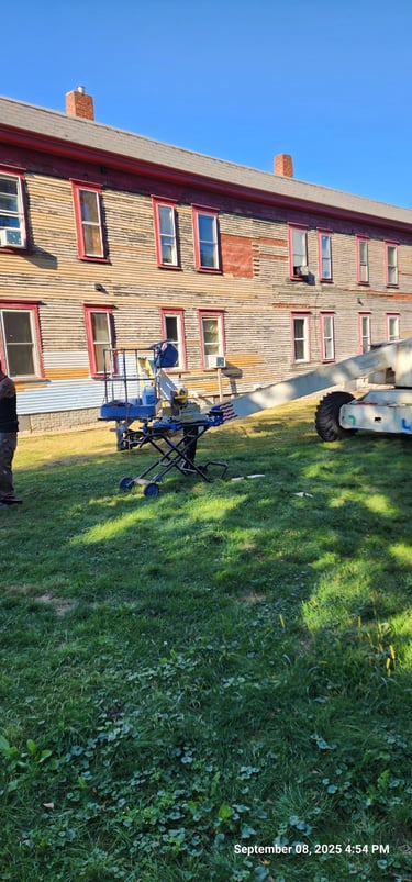 Exterior building restoration using a boom lift for siding repair and painting on an old wooden house.
