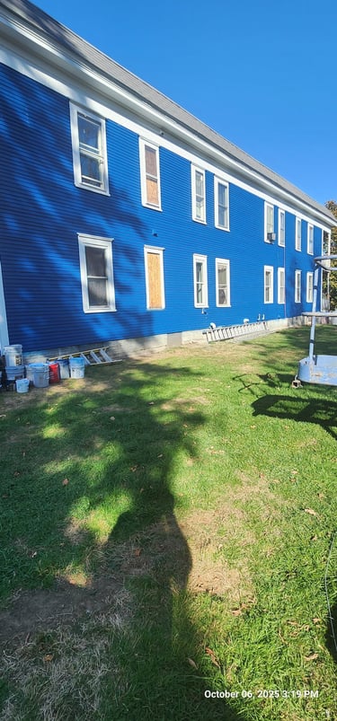 A two-story blue house with horizontal siding undergoing exterior painting and renovations on a sunny day.