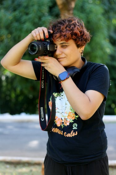 A young, short-haired person with a Canon DSLR camera taking a photo, with headphones around their neck and a blurry green