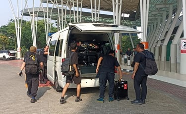 Lima orang laki-laki dewasa sedang menaikkan bagasi ke mobil Hiace warna putih.