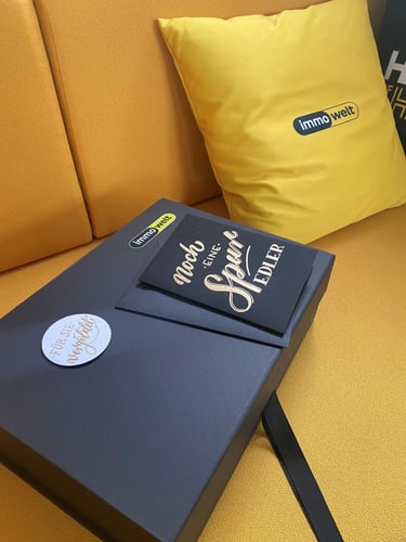 Elegant Immowelt corporate gift box and pillow on a yellow sofa with gold-lettered card.
