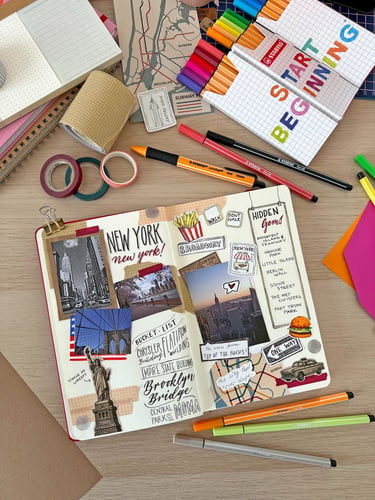 A colorful New York travel journal spread with stickers, photos, and sketches surrounded by Stabilo markers and stationery.