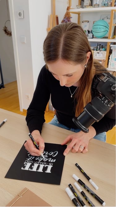 Artist using a white acrylic paint marker for hand lettering on black cardstock while filming.